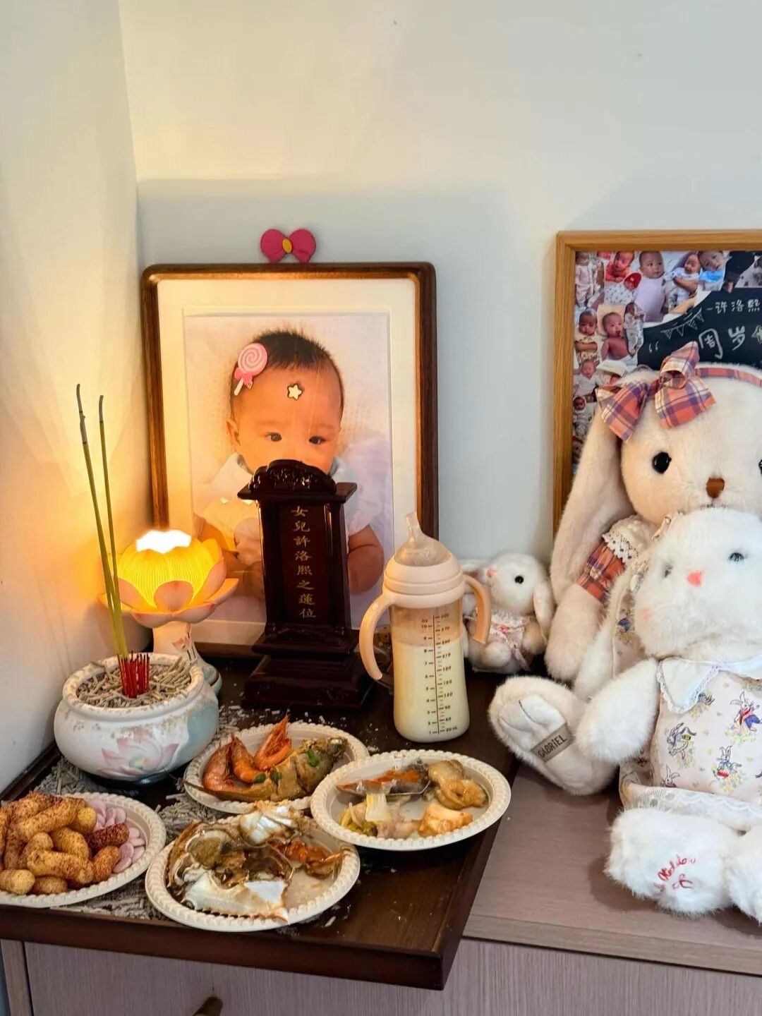 家中柜面上为小洛熙设的纪念角落：她的照片、刻着女兒許洛熙之蓮位的牌位、莲花灯与香炉、一瓶冲好的奶、几盘供菜、周岁纪念照拼贴和一圈兔子玩偶