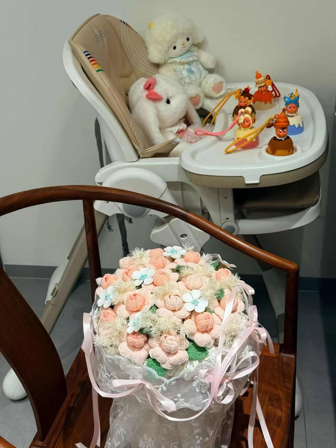 孩子的餐椅上坐着白色毛绒玩偶，托盘上摆着一排蛋糕造型的小挂件；椅前放着一束手工钩织的粉色花，用蕾丝与粉色缎带扎着