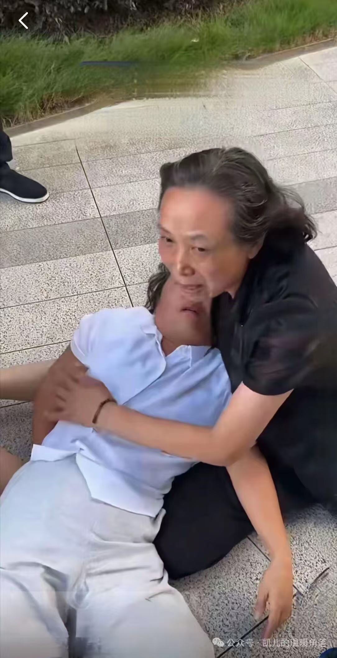 户外地砖路面上，一位年长女性抱着倒地的另一位女性，后者穿浅蓝上衣、白色长裤，闭目倒在地上；旁边可见他人的鞋与裤脚